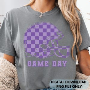 May include: Gray t-shirt featuring a purple checkered football helmet graphic. The words "GAME DAY" are printed below the helmet in purple. The bottom right corner of the shirt has the text "DIGITAL DOWNLOAD PNG FILE ONLY".