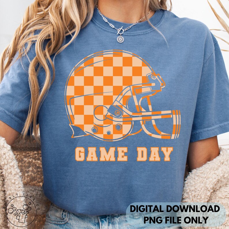 May include: Blue t-shirt with an orange and tan checkered football helmet graphic. The words "GAME DAY" are printed in orange below the helmet. The bottom right corner of the shirt has the text "DIGITAL DOWNLOAD PNG FILE ONLY".