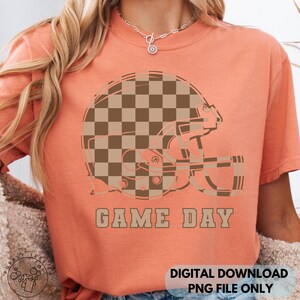 May include: A peach-colored t-shirt featuring a football helmet graphic with a brown and tan checkerboard pattern. The words "GAME DAY" are printed below the helmet in a matching brown color. The shirt is a digital download.