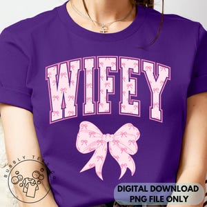 May include: A purple t-shirt with the word "WIFEY" in pink block letters filled with small pink bows. A large pink bow graphic is below the text. The shirt has rolled-up sleeves and a crew neck.