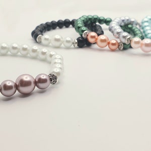 May include: A set of six pearl bracelets with different colored beads. The bracelets are made with white pearls, black lava beads, green beads, light blue beads, and peach beads. Each bracelet has a silver charm with rhinestones.