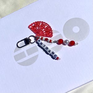 Peut inclure: Un porte-clés avec un charme en forme d'éventail rouge perlé avec le mot "ARIRANG" épelé en perles de lettres blanches. Le porte-clés comporte également des perles rouges et blanches, une perle et un fermoir argenté.