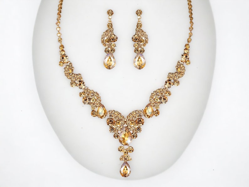 May include: A gold-toned jewelry set featuring a necklace and matching earrings. The necklace has a central heart-shaped design with dangling teardrop-shaped pendants. The earrings are drop style, with a similar design. The set is adorned with sparkling crystals.
