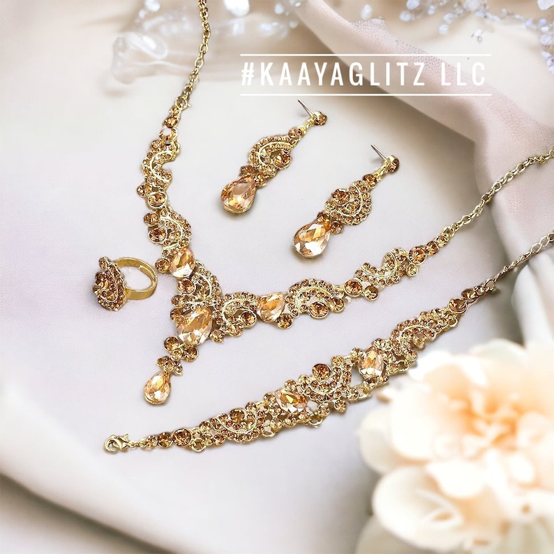 May include: A gold-toned jewelry set featuring a necklace, bracelet, earrings, and ring. The set is adorned with amber-colored gemstones and intricate detailing. The text "#KAAYAGLITZ LLC" is visible in the image.