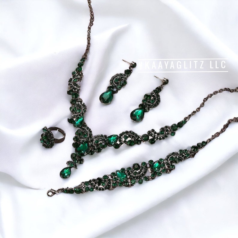 May include: A green emerald jewelry set with a necklace, bracelet, earrings, and ring. The jewelry is made of dark metal and features a floral design with teardrop-shaped stones.