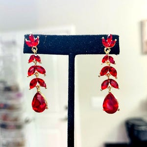 May include: A pair of gold-tone drop earrings featuring a vertical arrangement of red faceted stones. Each earring consists of three marquise-shaped stones followed by a single teardrop-shaped stone at the base, all connected by small gold metal links.