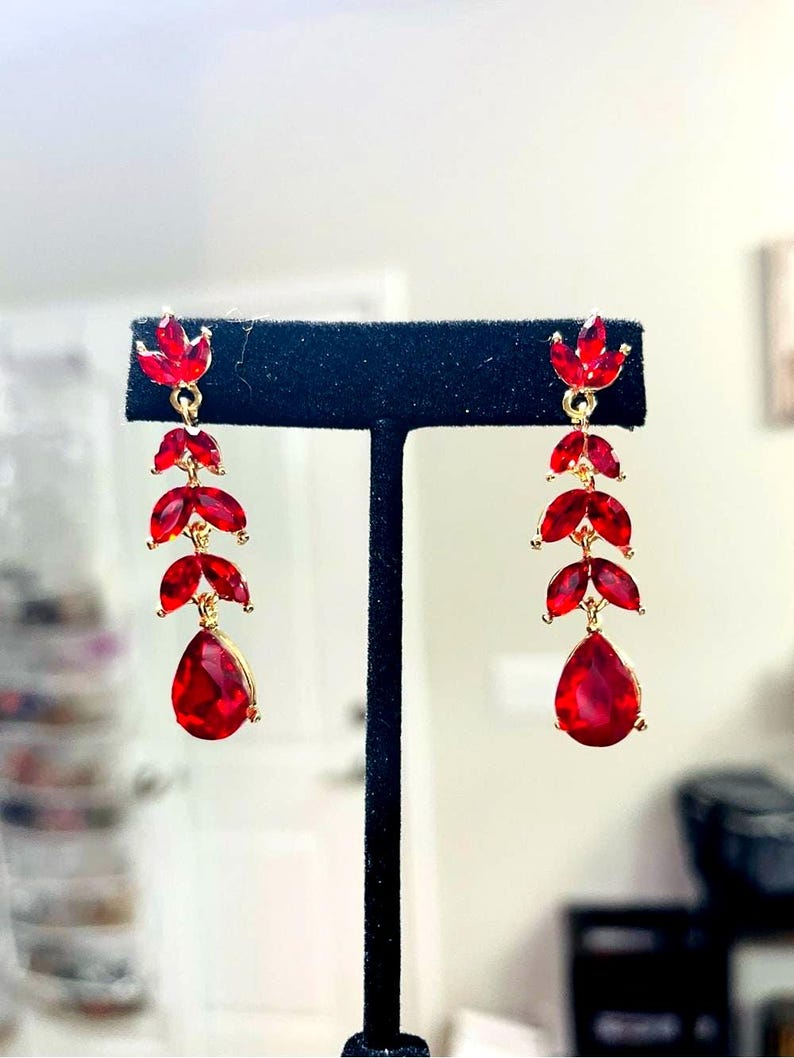 May include: A pair of gold-tone drop earrings featuring a vertical arrangement of red faceted stones. Each earring consists of three marquise-shaped stones followed by a single teardrop-shaped stone at the base, all connected by small gold metal links.