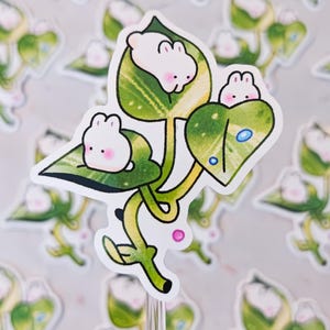 May include: A sticker featuring a whimsical illustration of three white bunnies nestled in green leaves. The leaves have a detailed, veined pattern, and the bunnies have pink cheeks. The sticker has a clear, glossy finish.