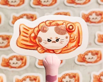 May include: A sticker featuring a cartoon cat wearing a taiyaki, a Japanese fish-shaped pastry. The cat has brown and white fur and is looking at the viewer with a grumpy expression. The taiyaki is orange and has a red bean filling.