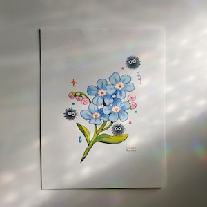 May include: Watercolour painting of a blue forget-me-not flower with three black and white Totoro characters. The painting is signed "Нокеи Рокеи" in the bottom right corner.
