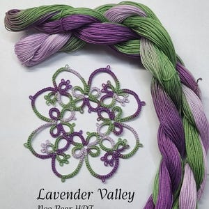 May include: A close-up of a delicate, hand-crafted lace doily in shades of green and purple. The doily is called "Lavender Valley" and is designed by "Noo Bear HDT".  Two skeins of thread in shades of green, purple, and lavender are braided together.
