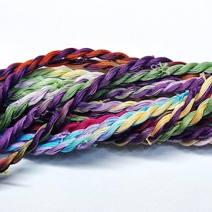 Puede incluir: Una colección de madejas de hilo de bordar de colores vivos en tonos de verde, morado, azul, amarillo y rojo. Las madejas están retorcidas y dispuestas en forma de abanico. Las madejas están etiquetadas "Outback 100% Seda - 10m Madeja - Hecho en Francia".