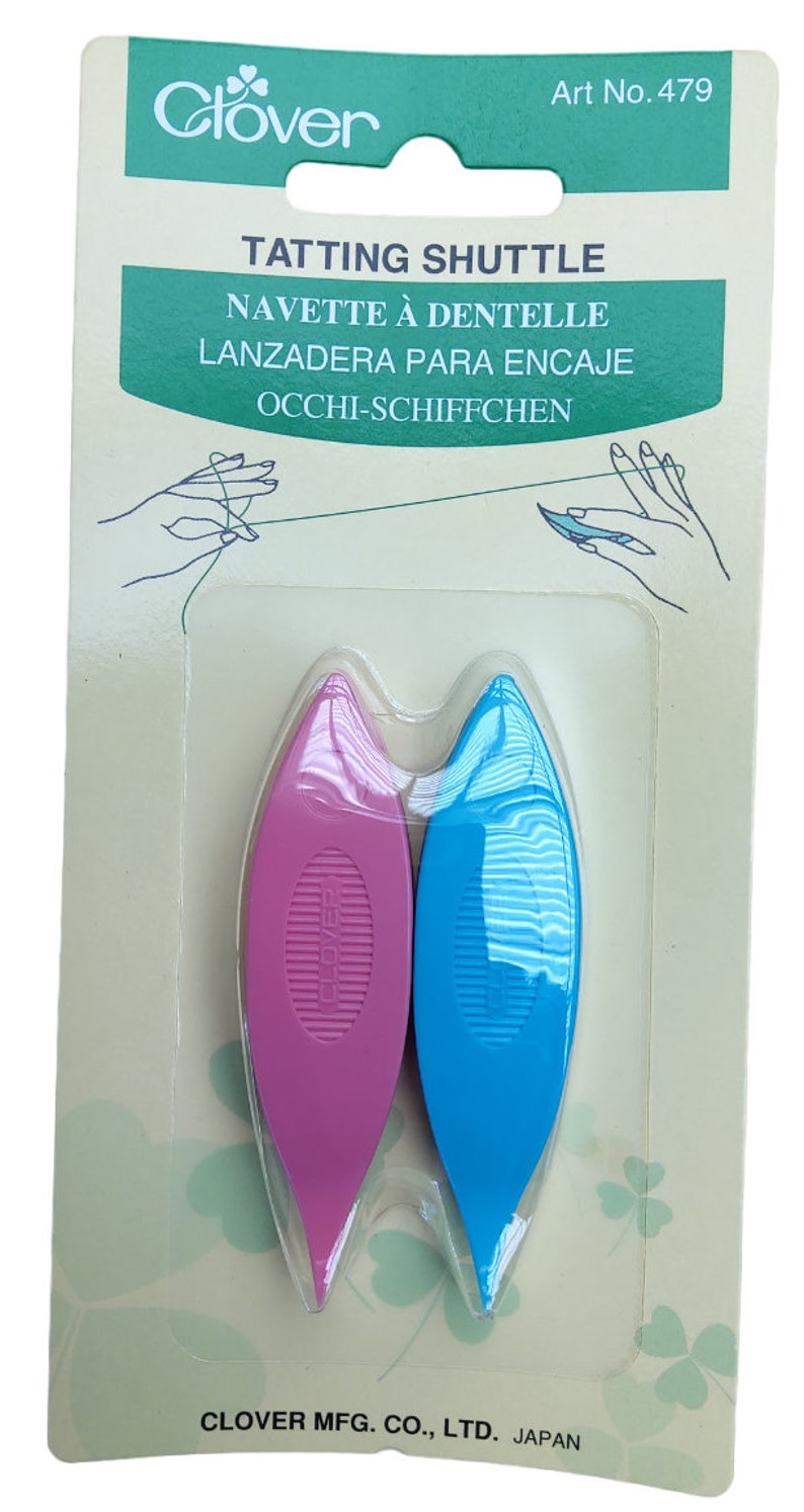 Op de afbeelding: Twee plastic tatting shuttles, &eacute;&eacute;n roze en &eacute;&eacute;n blauw, verpakt op een witte kaart met groene klaverontwerpen. De kaart vermeldt "Clover Tatting Shuttle" en "Art No. 479".