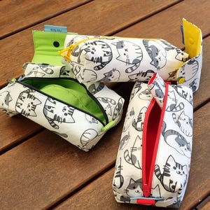 May include: Several white pencil cases with a repeating pattern of gray cat illustrations. One case is open, revealing a green interior. Another has a yellow zipper and a yellow interior. A third case has a red zipper.