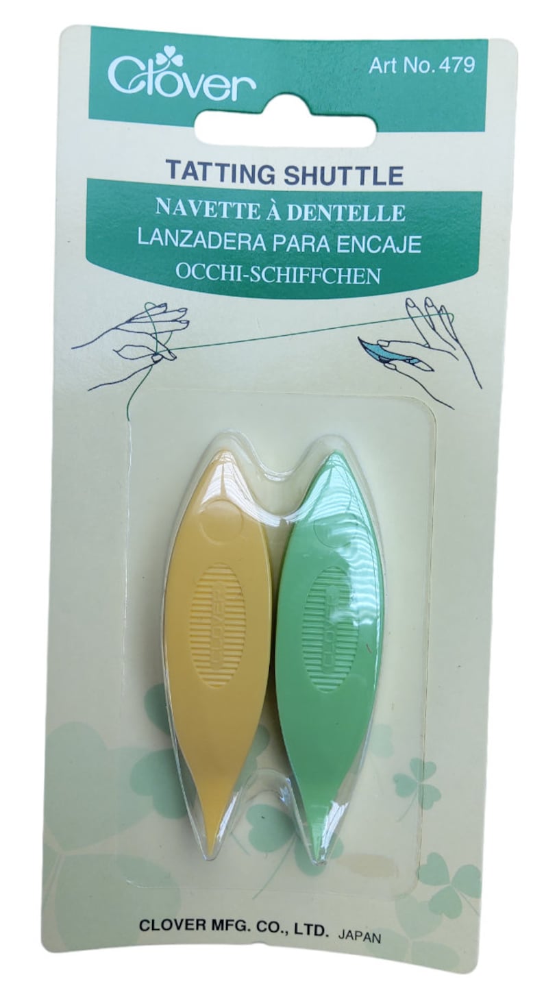 Op de afbeelding: Een verpakking met twee gele en groene plastic tatting shuttles. De verpakking is gelabeld "Clover Tatting Shuttle" en bevat de woorden "Navette &agrave; dentelle", "Lanzadera para encaje" en "Occhi-Schiffchen".