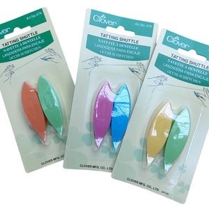 Op de afbeelding: Drie verpakkingen met Clover-tattingshuttles. Elke verpakking bevat twee shuttles in verschillende kleuren. De shuttles zijn gemaakt van plastic en hebben een gebogen vorm. De verpakkingen zijn gelabeld met de productnaam, het merk en het artikelnummer.
