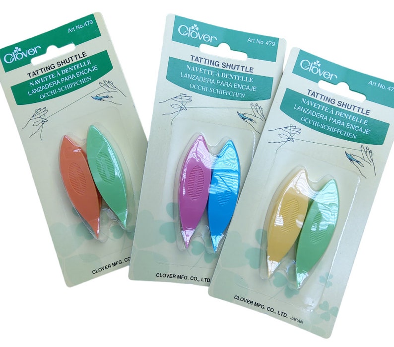 Op de afbeelding: Drie verpakkingen met Clover-tattingshuttles. Elke verpakking bevat twee shuttles in verschillende kleuren. De shuttles zijn gemaakt van plastic en hebben een gebogen vorm. De verpakkingen zijn gelabeld met de productnaam, het merk en het artikelnummer.