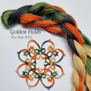 Può includere: Primo piano di un centrino tatting fatto a mano con un motivo floreale, con filo arancione, verde e blu navy. Un gomitolo di filo da ricamo variegato in colori simili è sullo sfondo. Il testo "Golden Fields" è visibile.