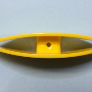 Op de afbeelding: Een geel plastic gereedschap met een gebogen handvat en een klein gaatje in het midden. Het gereedschap is bedoeld om te helpen met naaien of knutselen.