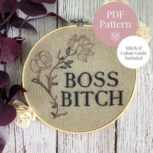 May include: A brown embroidery hoop with a beige linen fabric inside. The fabric has a black embroidered design of a floral stem with the words "BOSS BITCH" in black lettering. The hoop is on a white wooden surface.