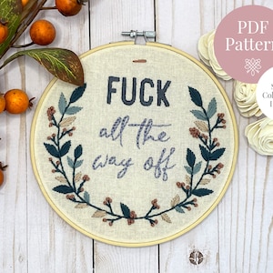 May include: Embroidered hoop art with the phrase "F*ck all the way off" surrounded by a wreath of green leaves and brown berries. The hoop is on a white wooden background.