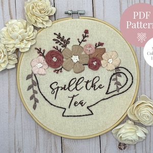May include: Embroidered hoop art with a teacup and the text "Spill the Tea". The teacup is filled with pink, brown, and white flowers. The hoop is on a wooden background.