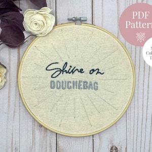 May include: A beige embroidery hoop with the text "Shine on Douchebag" stitched in black thread. The hoop is on a white wooden surface with a pink PDF pattern sign.