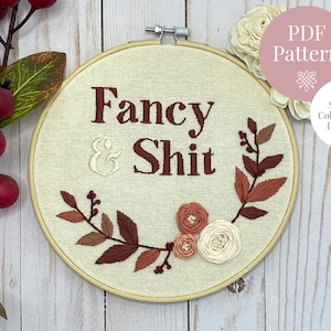 May include: Embroidered hoop art with the words "Fancy & Shit" in dark brown thread. The design includes a floral and leaf border in shades of brown and cream. The text "PDF Pattern" and "Stitch & Colour Guide Included" are also visible.