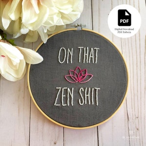May include: A round, dark gray embroidery hoop with the words "ON THAT ZEN SHIT" stitched in white thread. A pink lotus flower is embroidered below "ON THAT". The hoop is set against a light wood background, with a white flower in the upper left corner.