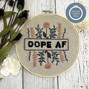 May include: Embroidered hoop art with the text "DOPE AF" in black, surrounded by floral embroidery in shades of blue, peach, and brown. The hoop is made of wood and the fabric is a neutral beige color. A "Finished Custom Hoop" label is in the upper right corner.
