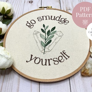 May include: Embroidery hoop with the words "go smudge yourself" stitched in brown thread. The design includes crystals and leaves in green and gray. The text "PDF Pattern" and "Stitch & Colour Guide Included" are also visible.