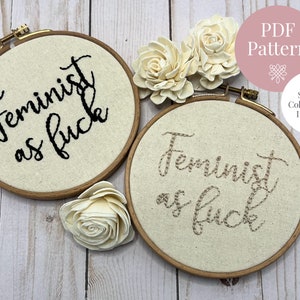 May include: Two round embroidery hoops with the text "Feminist as fuck" stitched in black and tan thread. The hoops are set against a light background with white flowers and the words "PDF Pattern" and "Stitch & Colour Guide Included".