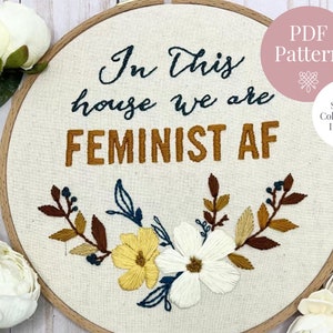 May include: Embroidered hoop art with the text "In this house we are FEMINIST AF" in brown and black lettering. The hoop is decorated with a floral design in shades of yellow, white, brown, and blue.