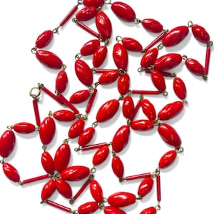 Op de afbeelding: Een opvallende rode ketting met ovale en cilindrische kralen. De kralen zijn verbonden door goudkleurige metalen schakels, wat een ingewikkeld patroon creëert. De ketting wordt gepresenteerd op een witte achtergrond.