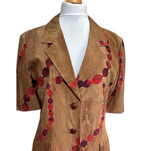 Op de afbeelding: Een bruin jasje met korte mouwen, met een herhalend patroon van rode en paarse ovale vormen. De blazer heeft twee voorzakken en drie bruine knopen. De stof heeft een subtiel textuurontwerp.