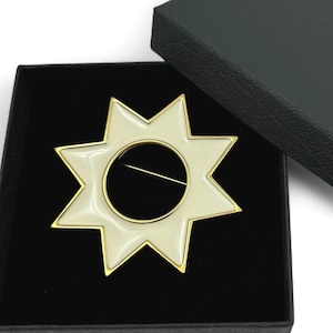 Könnte beinhalten: Sternförmige Brosche mit cremefarbener Emaille und goldfarbener Umrandung, präsentiert in einer schwarzen Geschenkbox. Die Brosche hat ein zentrales kreisförmiges Design und einen Nadelverschluss. Die Box ist geöffnet und zeigt den Schmuck.