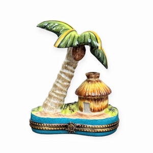 Puede incluir: Una figurita de porcelana de una palmera con una cabaña de paja debajo. La palmera tiene hojas verdes y un tronco marrón. La cabaña es marrón y tiene un techo de paja. La figurita está sobre una base azul con un diseño de sol.
