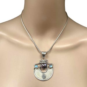 May include: A silver pendant necklace with a textured, half-moon pendant featuring a spiral design. The pendant has a central dark purple stone and two light blue stones. The necklace has a silver chain.