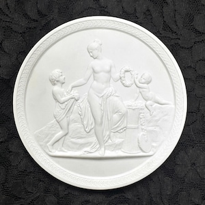 May include: A white circular bas-relief sculpture featuring classical figures. A central female figure is flanked by two smaller figures, one holding a wreath. The relief has a detailed border and is set against a dark background.
