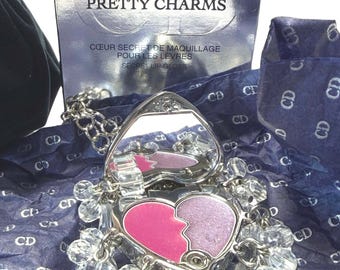 May include: A silver heart-shaped compact with a mirror and two shades of pink eyeshadow. The compact is adorned with clear crystal beads and a chain. The box behind it reads "DIOR PRETTY CHARMS" and "SECRET LIP GLOSS."