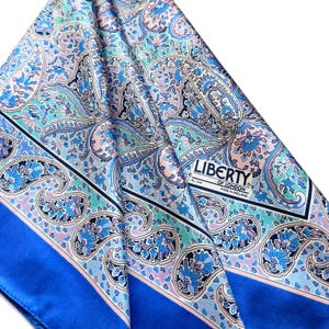 May include: A square silk scarf featuring a detailed paisley pattern in shades of blue, light purple, and soft pink. The fabric includes a solid blue border and a white label that reads LIBERTY OF LONDON ALL SILK MADE IN ENGLAND in black text.