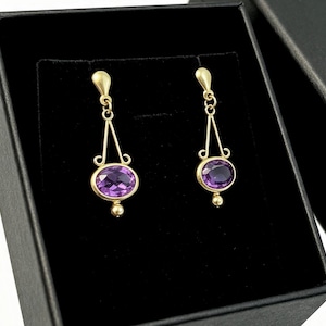 May include: A pair of gold drop earrings featuring oval-cut purple amethyst gemstones. Each earring has a delicate gold wire frame with a small gold bead at the base, suspended from a simple gold hook, presented in a black jewellery box.