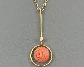 Può includere: Una collana con pendente dorato con una rosa di corallo scolpita all'interno di un cerchio dorato. Un piccolo diamante è incastonato sulla barra verticale. La collana pende da una delicata catena d'oro, creando un gioiello elegante.