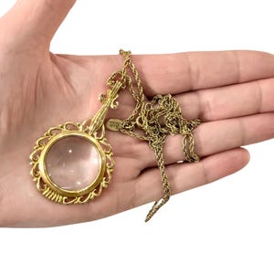Op de afbeelding: Een goudkleurige vergrootglas-hanger aan een bijpassende goudkleurige touwketting. De ronde lens is gevat in een sierlijk metalen frame in filigraanstijl met een decoratief handvat dat naar boven uitsteekt en een klein metalen label bij de ketting.