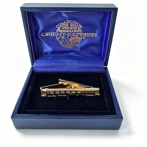 May include: A gold-toned tie bar shaped like a vintage train carriage, presented in a dark blue hinged display box. The interior lid features the gold-printed text COLLECTION VENICE SIMPLON ORIENT-EXPRESS with a circular logo design.