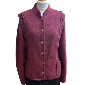 Könnte beinhalten: Burgunderfarbene Wolljacke mit Stehkragen und silberfarbenen Knöpfen. Die Jacke hat einen strukturierten Stoff, geflochtene Schulterakzente und zwei Vordertaschen. Die Ärmel sind lang und die Jacke ist zugeknöpft.