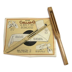 May include: A light brown wooden collar tool alongside its original instructional paper packaging. The paper features the text ACME COLLAR-Q, A Boon to men, and includes illustrations demonstrating how to insert a collar into the wooden device.