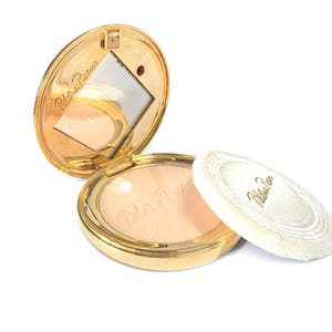 May include: A gold-toned compact with a mirror and a powder puff. The compact is open, revealing the powder inside. The powder puff is white with a gold-colored border. The words "Paloma Picasso" are printed on the compact and puff.
