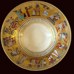 May include: A decorative gold-colored plate with an Egyptian theme. The plate features a central cream-colored bowl surrounded by a gold border with detailed illustrations of Egyptian figures and hieroglyphs. The rim has a repeating pattern of blue and gold accents.