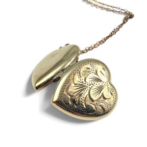 Könnte beinhalten: Ein herzförmiger Medaillon-Anhänger in Goldoptik an einer passenden Kette. Das Medaillon ist geöffnet und zeigt eine glatte Innenseite mit der Prägung ROLLED GOLD. Die Außenseite des Medaillons ist mit einem floralen Gravurmuster versehen.